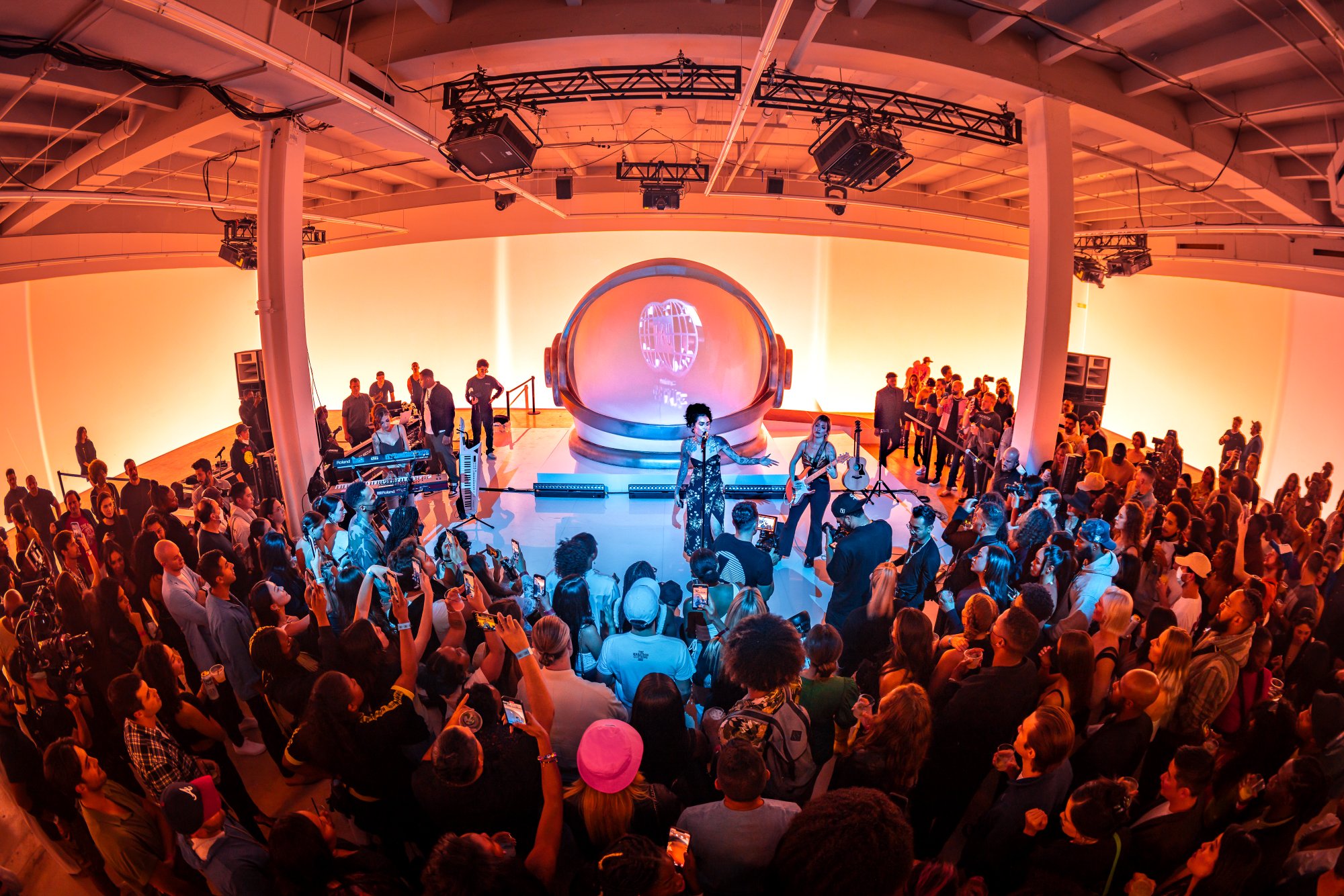 A packed crowd around a stage with a giant astronaut-helmet sculpture, warm light