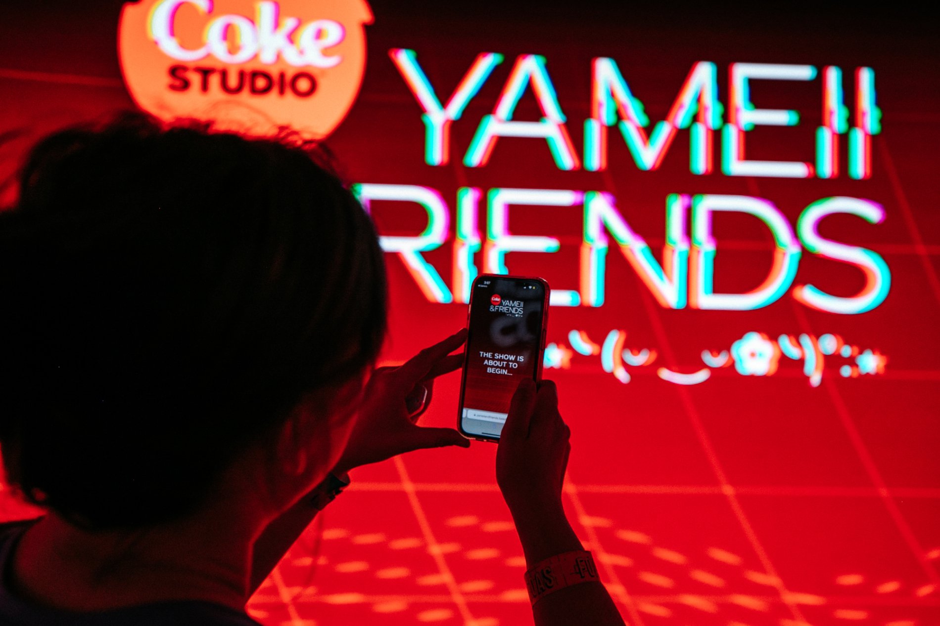 A guest holds up a phone before the glowing Coke Studio Yameii & Friends wall