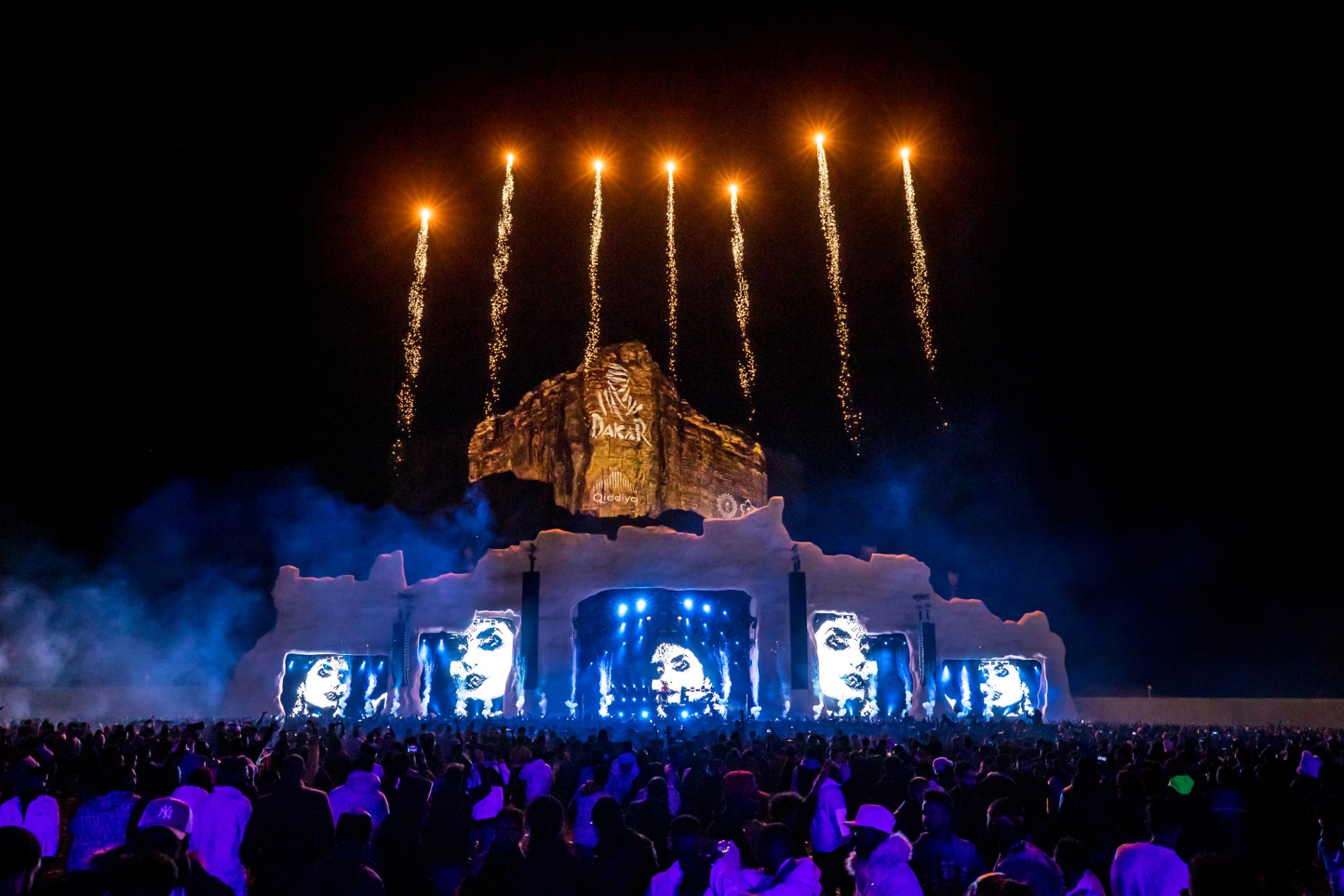 A face projected across the full cliff in blue, fireworks rising above the crowd