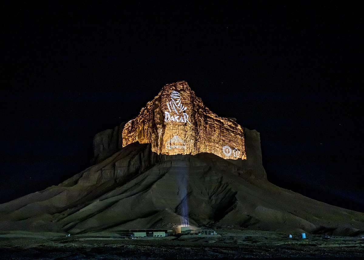 The Dakar logo projected onto Tuwaiq Mountain at night, a single beam climbing the rock