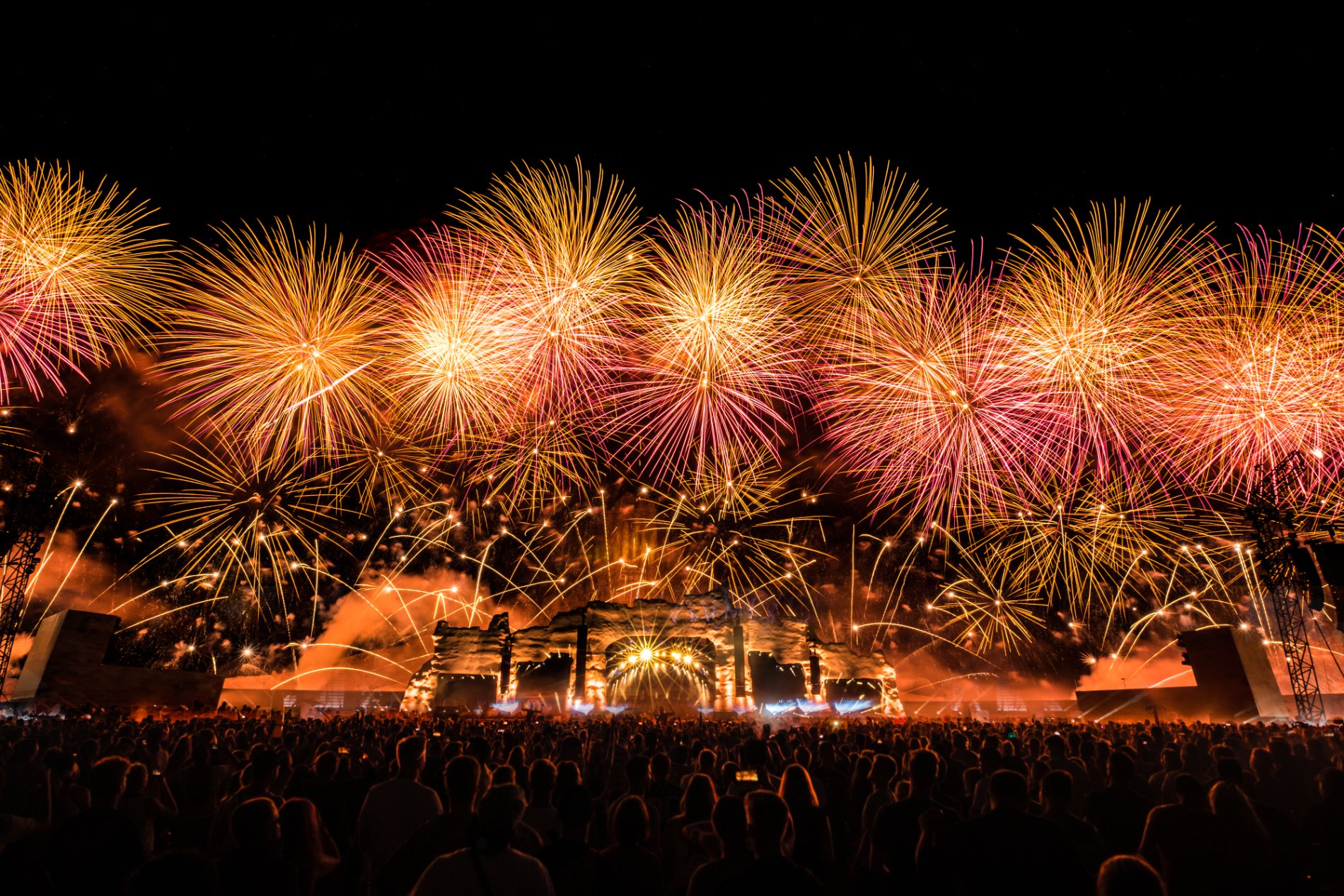 A wall of gold fireworks fills the sky over the lit rock stage and the desert crowd