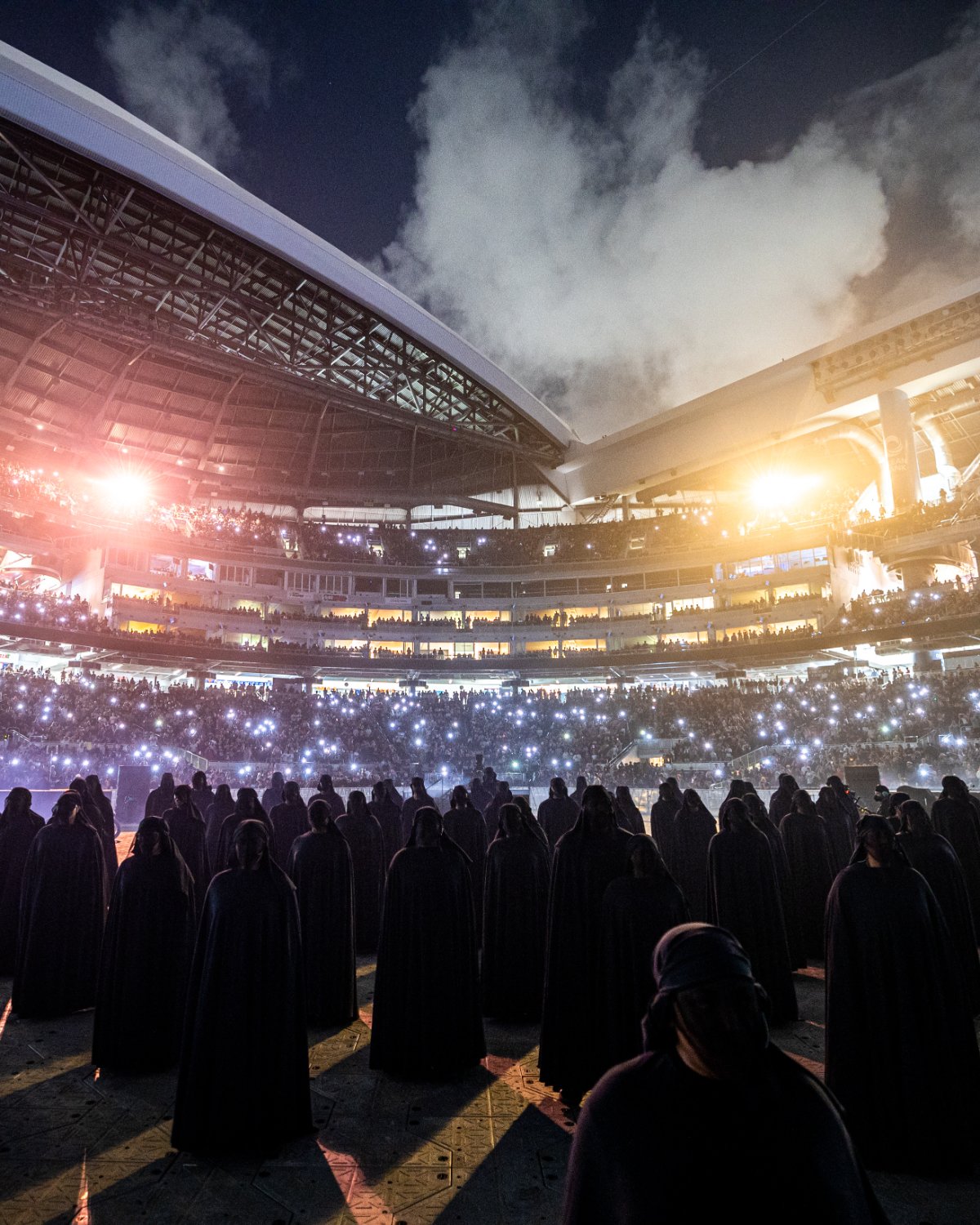Cloaked performers fill the ballpark floor under an open sky and stadium light