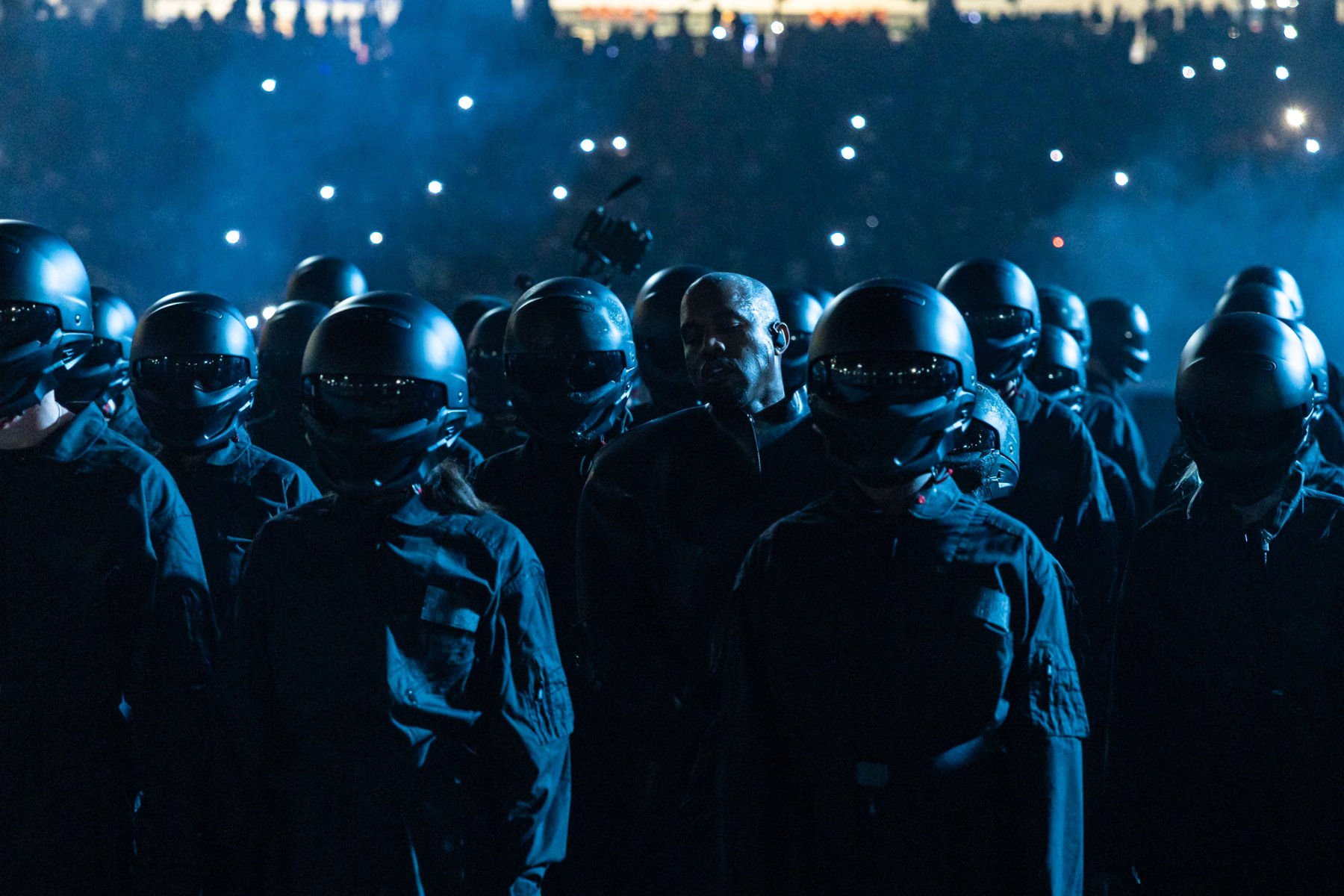 Kanye West among a formation of helmeted performers in blue light