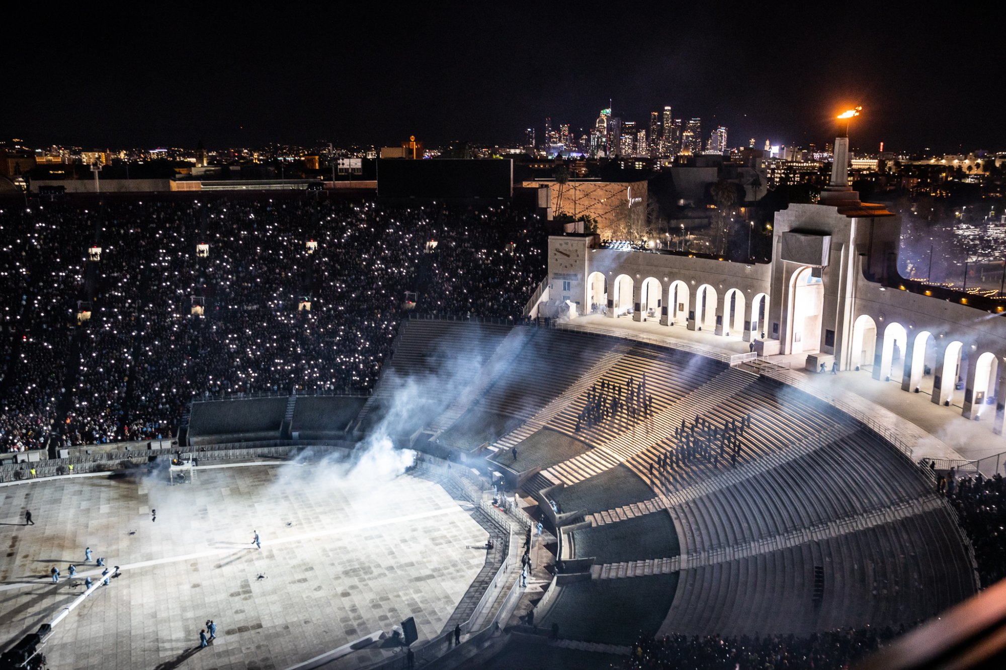 Aerial of the Coliseum, the LA skyline behind, the full crowd lit by phones