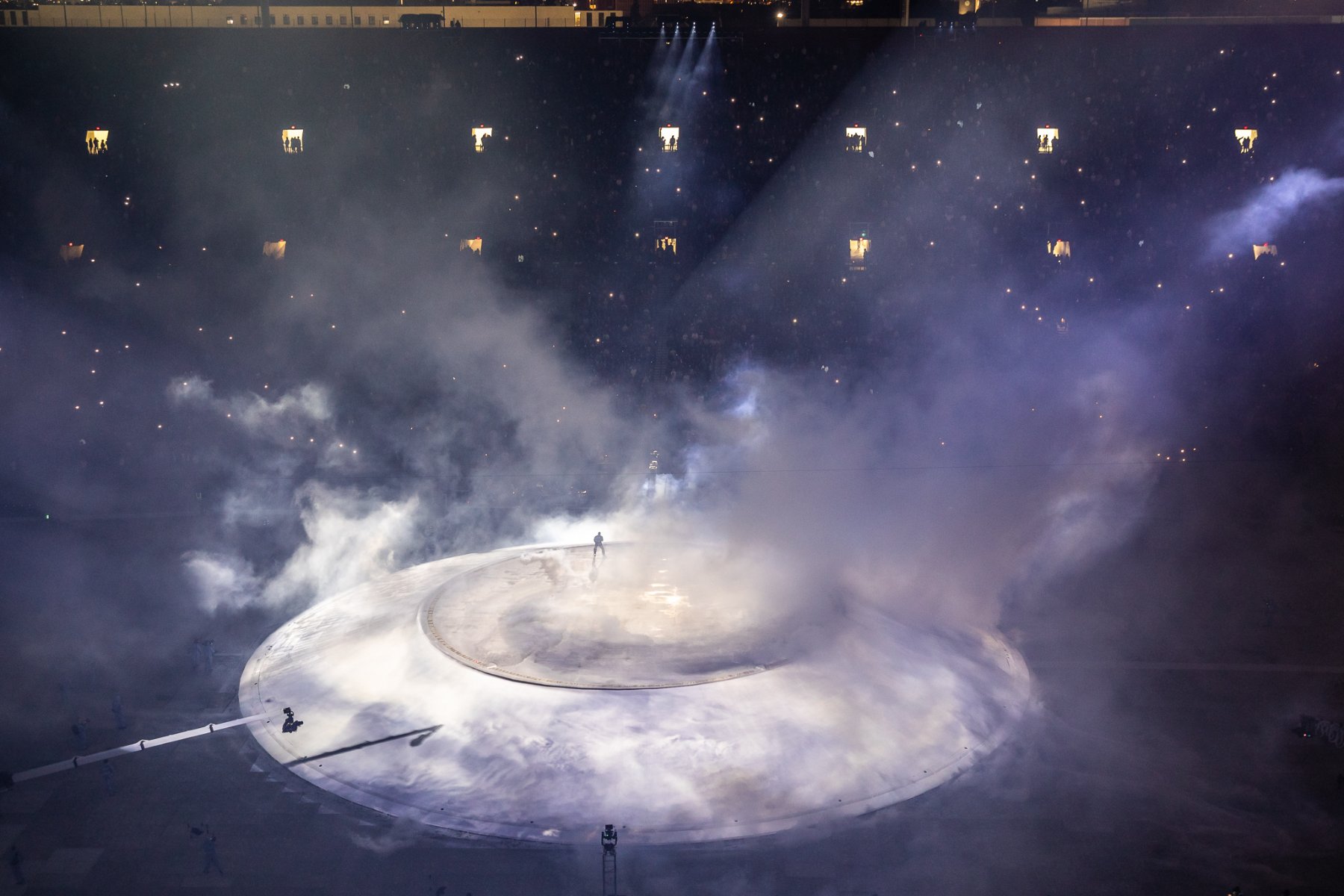 Overhead of the round Mound, a lone figure in a spotlight through smoke