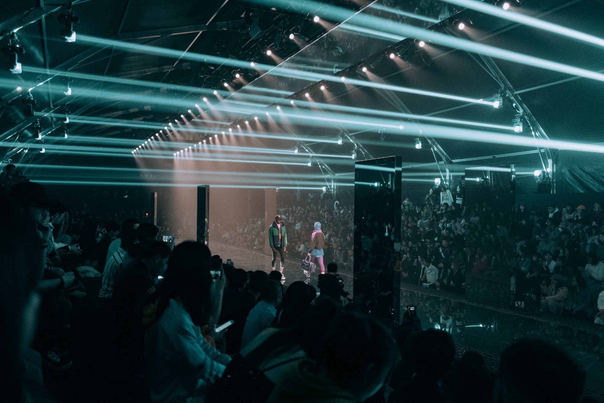 Wide view of the runway, teal beams raking across the full crowd