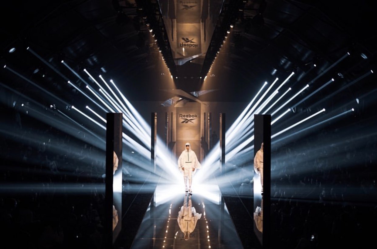A lone model walks a mirrored runway between monoliths under radial beams of light