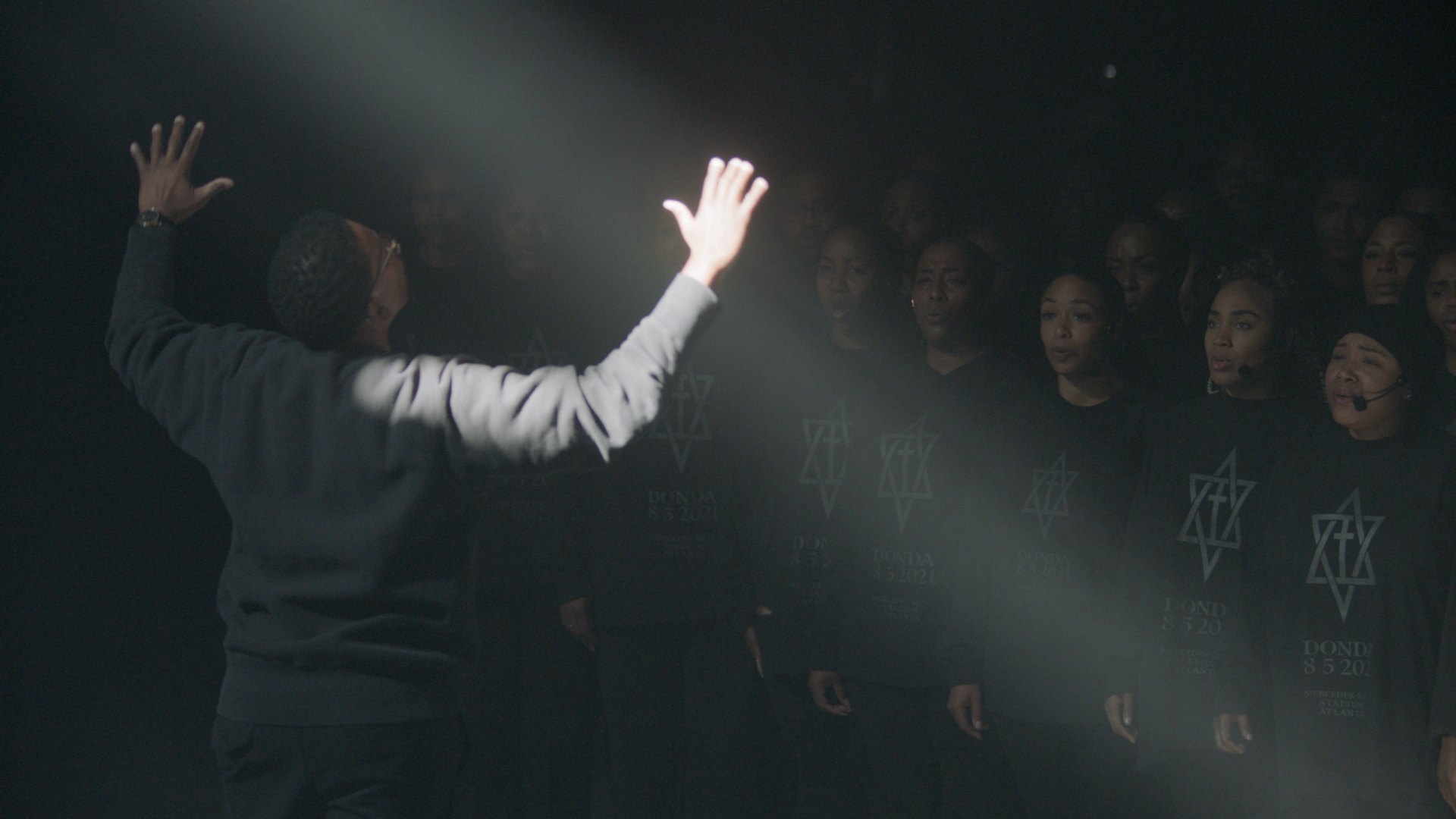 The conductor seen from behind, arms raised, the choir in DONDA shirts
